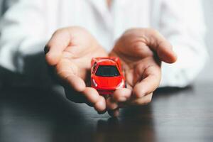 företag kvinnas hand skyddande röd leksak bil på skrivbord. planera till klara av transport finansiera kostar. begrepp av bil försäkring företag, sparande köpa - försäljning med beskatta och lån för ny bil. foto