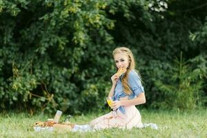 en leende skön flicka innehar en färsk bulle i henne händer och sitter på en filt i de öppen luft i de parkera. sommar picknick i de parkera foto