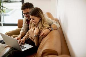 ungt par som använder bärbara datorn tillsammans medan du sitter på soffan hemma foto