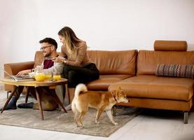 ung kvinna och ung man som använder bärbar dator för onlinebetalning medan de sitter vid soffan med sin shiba inu-hund hemma