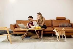 ung kvinna och ung man som använder bärbar dator för onlinebetalning medan de sitter vid soffan med sin shiba inu-hund hemma