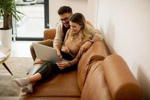 ungt par som använder bärbara datorn tillsammans medan du sitter på soffan hemma foto