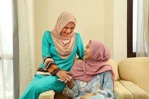 två ung asiatisk malaysiska muslim kvinna bär slöja på Hem kontor studerande Sammanträde på soffa prata blanda leende Lycklig foto