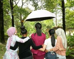 grupp av kvinna vänner malaysiska kinesisk indisk asiatisk utomhus- parkera natur gående under ett paraply säkra säker enhet händer på axel och midja foto