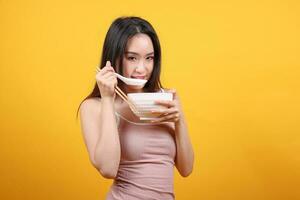 skön ung söder öst asiatisk kvinna innehav chines tömma matpinne kinesisk soppa sked skål redskap låtsas verkande Framställ ser äta smak lukt utfodra erbjudande tillfredsställelse smaskigt gul orange bakgrund foto