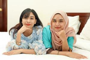 två ung asiatisk malaysiska muslim kvinna bär slöja baju kurung klänning på Hem på säng utgör Lycklig självsäker se på kamera hand på haka ansikte foto