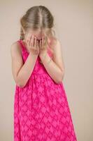 liten flicka i en rosa klänning på en beige bakgrund omslag henne ansikte med henne händer foto