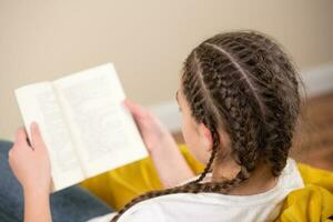Tonårs flicka med flätor läsning bok på gul bönpåse stol foto