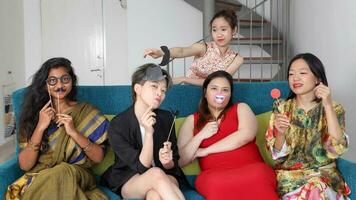 ung asiatisk kvinna grupp prata sjunga framställning rolig hånfull ansikten uttryck selfie porträtt papper rekvisita mustasch hatt rör näsa hår på blå levande rum soffa sitta foto