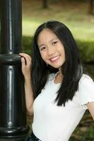 ett asiatisk kinesisk kvinna Framställ leende se framåt- mot kamera självsäker utomhus- grön parkera foto