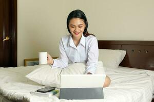 ung skön modern malaysiska kvinna sitta på säng arbete studie uppkopplad i sovrum bärbar dator flik kaffe kopp bok foto