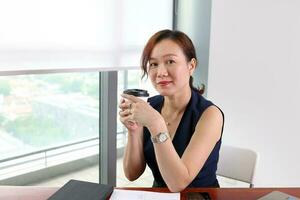 skön attraktiv mitten ålder sydöst asiatisk kvinna Hem kontor arbete studie sitta på tabell dryck kaffe kopp se framåt- främre urban dagsljus fönster bakgrund urban dagsljus fönster bakgrund foto