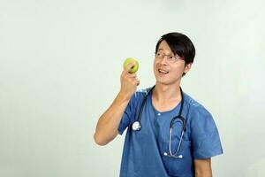 ung asiatisk manlig läkare bär förkläde enhetlig tunika stetoskop innehav pekande som visar äter friska grön äpple foto