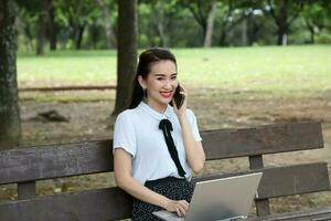 ung asiatisk kinesisk kvinna utomhus- på parkera bänk använda sig av bärbar dator dator tror läsa se Lycklig undra leende främre prata på telefon foto