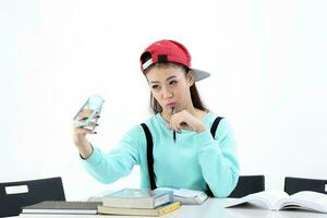 yong asiatisk ungdom flicka i tillfällig klänning keps sitta på tabell bok anteckningsbok penna selfie smart telefon på vit bakgrund foto
