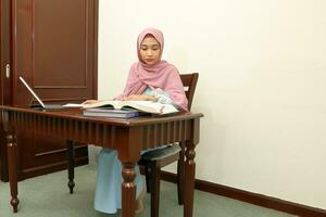 ung asiatisk malaysiska muslim kvinna bär slöja på Hem kontor studerande sitta på tabell dator läsa bok fil papper studie arbete foto