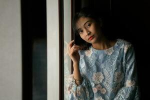 ung asiatisk malaysiska kvinna bär baju kurung klänning på Hem i främre fönster humör ljus på ansikte mörk bakgrund fredlig innehåll lugna skytte lugn foto