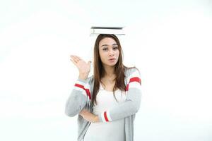 yong asiatisk ungdom flicka i tillfällig klänning balansering bok på huvud se på kamera hand i luft uttryck rolig ansikte på vit bakgrund foto