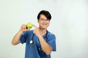 ung asiatisk manlig läkare bär förkläde enhetlig tunika stetoskop innehav pekande som visar äter friska grön äpple foto