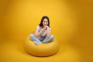 skön ung söder öst asiatisk kvinna sitter på en gul bönpåse sittplats orange gul Färg bakgrund utgör mode stil elegant skönhet humör uttryck resten koppla av leende foto