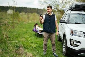 man i ärmlös väst håll telefon mot sUV bil med tak kuggstång låda, familj på picknick. foto