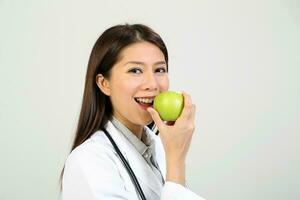 ung asiatisk kvinna läkare bär förkläde enhetlig tunika stetoskop innehav pekande som visar äter friska grön äpple foto