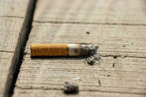 brinnande cigarett stänga upp på en bruten trä- yta. cigarett på en trä- yta stänga upp. ohälsosam rökning vana och cigarr missbruk. nikotin missbruk begrepp med en bränd cigarett och aska. foto