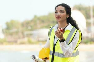 kvinna ingenjör med Hardhat med petrokemiska fabrik bakgrund. asiatisk kvinna innehav läsplatta, planen och walkie talkie. foto