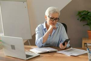 självsäker eleganta europeisk mitten åldrig senior kvinna använder sig av smartphone på arbetsplats. eleganta äldre mogna 60s grå håriga lady affärskvinna med cell telefon i kontor. chef ledare använder sig av internet appar. foto