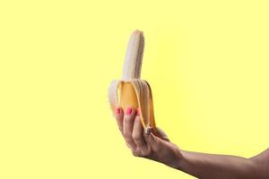 närbild en kvinna handmålade röda naglar som håller en mogen banan isolerad på gul pastellfärgad bakgrund foto