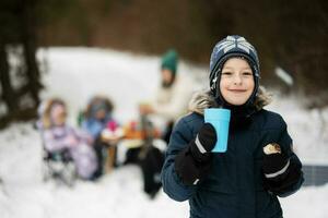 pojke med kopp av te på hand mot hans familj i vinter- skog utgifterna tid tillsammans på en picknick. foto