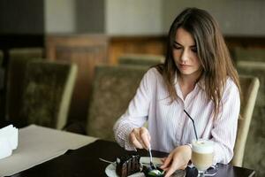 ung skön brunett kvinna sitta i kaffe affär Kafé restaurang inomhus och äta choklad tomte efterrätt kaka. foto