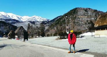 asiatisk turist man i röd täcka stående och bär solglasögon och gul sticka hatt Sammanträde på väg, gata eller väg med berg och himmel bakgrund i japansk by, japan. Asien resa, turism. foto