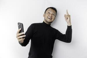 en porträtt av en Lycklig asiatisk man bär en svart skjorta och innehav hans telefon pekande upp på kopia Plats, isolerat förbi vit bakgrund foto