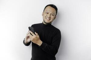 upphetsad asiatisk man bär svart skjorta leende medan innehav hans telefon, isolerat förbi vit bakgrund foto