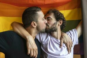 passionerad Gay par framställning ut med lgbtq flagga i de bakgrund. ung Gay par bindning förtjust inomhus. foto