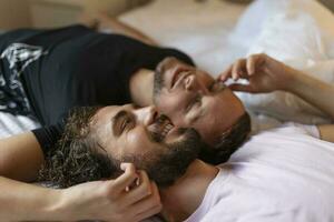 Gay par fattande varje Övrig med deras ögon stängd. två ung manlig älskande rörande deras ansikten tillsammans medan liggande i säng i de morgon. öm ung Gay par bindning på Hem. foto