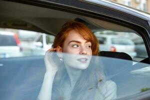 självsäker och skön. främre se av attraktiv ung röd hår kvinna i tillfällig ha på sig ser bort medan körning en bil under de dag. skön leende kvinna körning bil, Sammanträde i bil foto