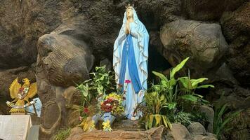 staty av helig jungfrulig mary i roman katolik kyrka, i de grotta av jungfrulig Mary, i en sten grotta kapell katolik kyrka med tropisk blommor runt om foto