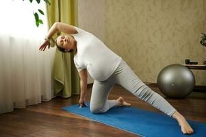 aktiva gravid kvinna med stor mage, stretching henne kropp medan praktiserande prenatal kondition på blå övning matta på Hem foto