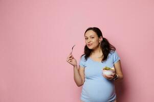glad gravid kvinna innehav skål med färsk organisk frukter, njuter friska livsstil och näring i graviditet tid foto