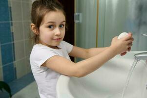 skön liten barn flicka leende på kamera, innehav tvål bar och tvättning henne händer, stående förbi handfat på Hem badrum foto