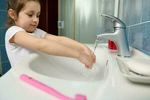 liten unge flicka tvättning händer under kran med löpning vatten på Hem badrum. rena händer, hygien och sanitär begrepp foto