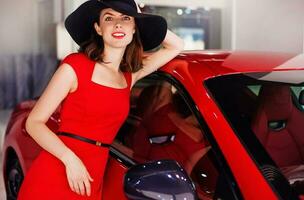 skön ung caucasian kvinna bär röd klänning stående med en röd bil foto