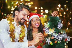 skön indisk par dekorera jul träd med lampor bokeh ett de bakgrund foto
