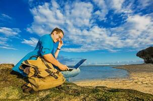 ung caucasian man Sammanträde på en strand arbetssätt på en bärbar dator med en väska liggande förbi hans sida foto