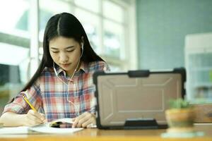ung asiatisk kvinna studie i främre av de bärbar dator dator och och använder sig av hörlurar på utanför platsen. foto