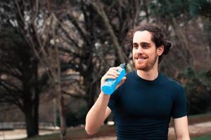 en ung atletisk man ler medan du håller en karbonatdrink i parken medan du gör kondition