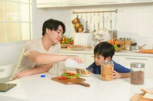 Lycklig ung asiatisk far och son äter friska mat i kök på Hem foto