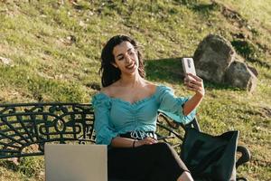 ung kvinna som tar en selfie på en bänk i parkens livsstilskoncept sommarstil foto